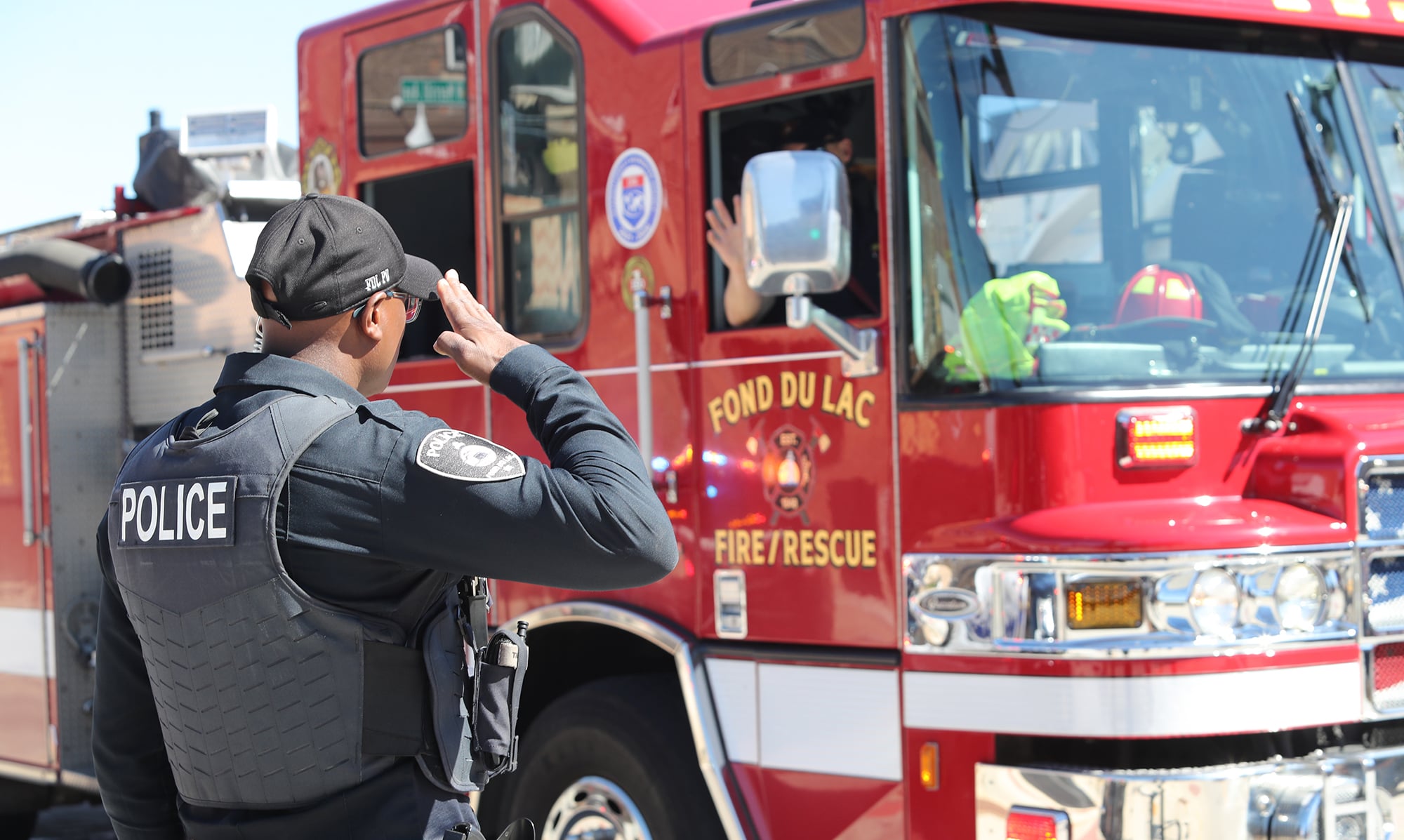 A Fond du Lac police officer in tactical gear stands at attention, saluting a passing Fond du Lac Fire/Rescue engine during a community event.